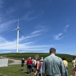 A group of people facing a wind turbine in an open field.